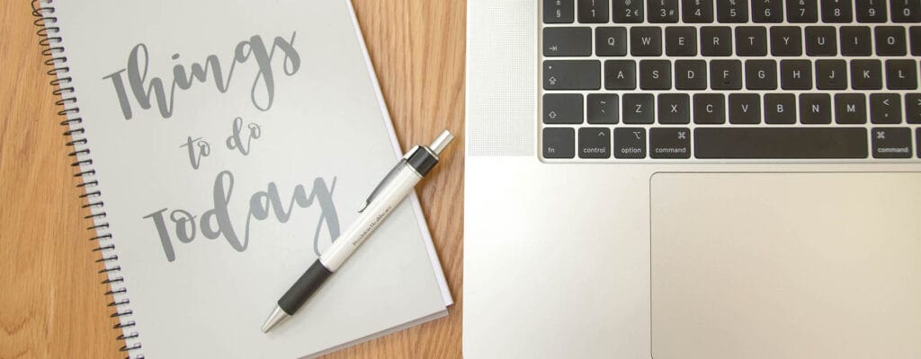 Flat Lay brand photograph showing a pen on top of a grey notebook, next to a silver and black business laptop keyboard on a wooden desk, The text on the notebook says “Things to do today”